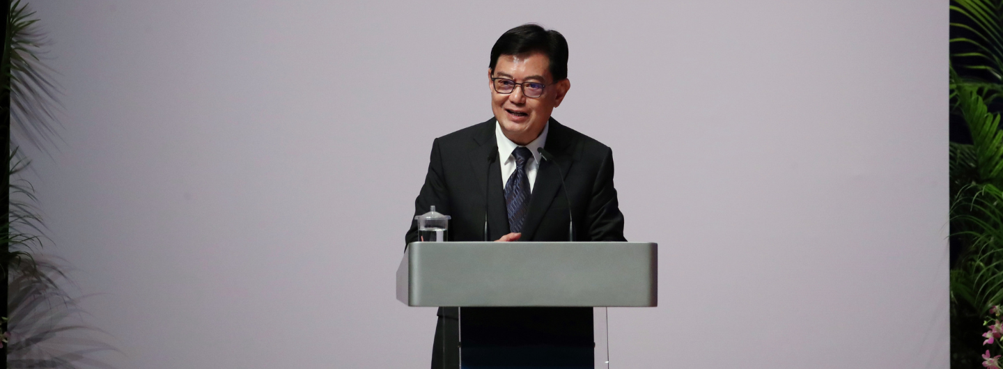Lawrence Wong at a podium, wearing a suit and tie, with a glass of water.